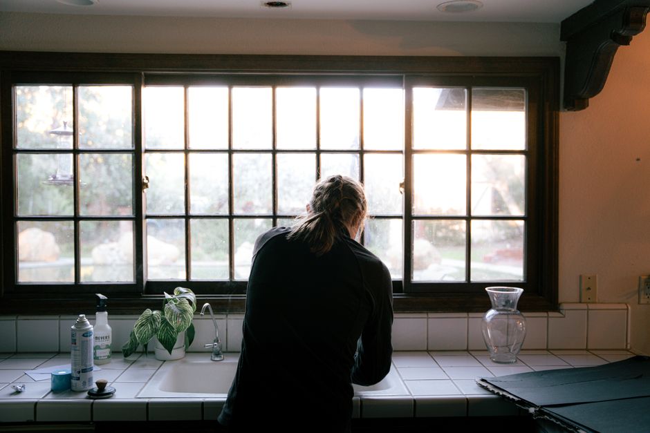 A woman washing dishes in a sunlit kitchen with large window view.